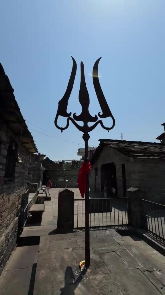 A tall black metal trishul against a blue sky beside stone temple buildings.