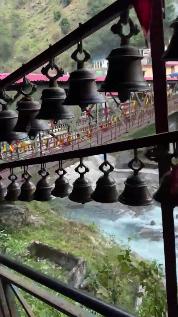 Rows of dark metal bells hanging along a railing, with a rushing river and green hillside below.